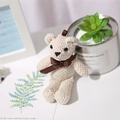 Beige knitted teddy bear plush toy with brown ribbon bow in a cozy home setting - White