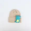 Nahaufnahme einer beigen Strickmütze für Babys mit einem blauen 3D-Dinosaurier-Aufnäher - Cartoon Dinosaurier / Beige