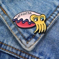 Macro detail of enamel cuttlefish pin with 'LET'S CUTTLE' text on blue denim jacket - Yellow Cuttlefish