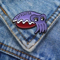 Detail of purple enamel cuttlefish pin with 'LET'S CUTTLE' text on blue denim jacket - Purple Cuttlefish
