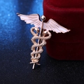 Dettaglio della spilla caduceo dorata con ali in smalto bianco su scatola di velluto rosso - Oro