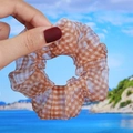 Close-up of brown plaid organza scrunchie held by hand against a blurred beach backdrop - Orange Plaid