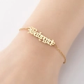 Vista macro de una pulsera dorada Babygirl con letra gótica en la muñeca de modelo - Dorado