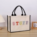 Close-up of colorful embroidered STUFF lettering on beige canvas tote bag with black handles - STUFF
