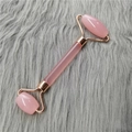 Dual-ended rose quartz facial roller with gold metal handle on a soft grey fur background - Pink Roller