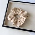 Macro detail of beige pleated fabric scrunchie on a display tray with logo label - Pleated Khaki