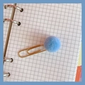 Detail of golden metal paperclip with a blue fuzzy pompom on a grid notebook page - #3 Blue Pom-pom Paperclip