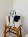 Detail of a large cream canvas tote bag with navy blue handles and knit gloves on a wooden stool - Off White
