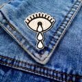 Detail of teary eye enamel pin on blue denim fabric background with gold metal edges - Tearing Eye