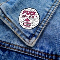 Détail d'un pin's émaillé rigolo avec texte rose épinglé sur une poche en jean bleu - Visage Texte Drôle