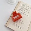 Square orange hair claw clip in glossy plastic shown on an open book background - Orange Red