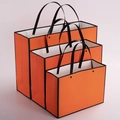 Three vibrant orange paper gift bags with black rivet reinforcement and ribbon handles - Orange With Black Border Rivet Style