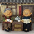 Elderly couple figurines sitting on a bench with traditional Chinese house background - 935-5 Reading Newspaper