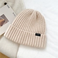 Close-up of thick ribbed knit wool beanie in cream white with black logo label - One Size / Beige