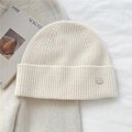 Close-up of rib-knitted beige wool beanie featuring a premium metallic brand logo tag - Cream