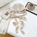 Close-up of oatmeal knitted bonnet hat with neck ties and white label on a cozy bed - Cream