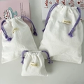 Detailliertes Set aus drei cremeweißen Krepp-Baumwollbeuteln mit lila Kordelzug - Groß / Weiß