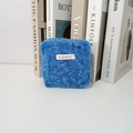 Detailed close-up of a blue plush square pouch with a mayrabbit woven label on white table - Blue