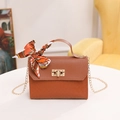 Brown woven crossbody bag with gold lock and orange scarf handle on a white table - Brown