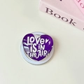 Nahaufnahme einer lila herzförmigen Handyhalterung mit der Aufschrift LOVE IS IN THE AIR - Magnetisch-Kristallglas Herz-Dunkellila Beschriftung