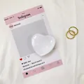 Flat lay of a transparent glass heart phone grip and two gold rings on a pink display card - White