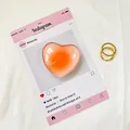 Close-up of translucent heart-shaped phone grip in soft pink with gold rings on fabric - Orange Dots