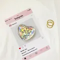 Dettaglio del supporto per telefono olografico a cuore e anelli dorati su tessuto bianco - Cuore Laser Olografico