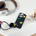 Custodia iPhone nera con cuori 3D, ciondolo a pompon e cinturino su scrivania moderna - Nero / iPhone 15