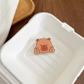 Close-up of a cute cartoon capybara acrylic phone grip with a winking face on a tray - Winking Peace Sign Capybara Style (17号)
