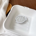 Close-up of a fish-shaped mirror phone grip with scales inside a white box - White