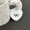 Detail of a white heart-shaped phone grip featuring a black bow design on a ceramic cup - White