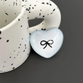 Close-up of a light blue heart-shaped phone grip with a black bow print on a ceramic cup - Light Blue