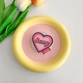Detail of a pink heart-shaped MagSafe phone grip with Bunny typography on a ceramic dish - Rose Red Bunny