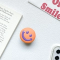 Detail of an orange collapsible phone grip with a purple smiley face on a white fabric - Orange