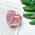 Detailweergave van de hartvormige glitter telefoonhouder met roze lovertjes op boekachtergrond - Gefacetteerde Hartstandaard - Zilverfolie - Transparant Rood