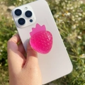 Nahaufnahme eines Handy-Sockels aus rosa Harz in Erdbeerform auf weißem Smartphone - Glänzende Rosenrote Erdbeere