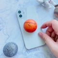 Orange planet-shaped expanding phone grip stand with textured surface on a white smartphone - Mars