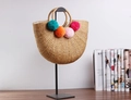 Dettaglio borsa a mano in paglia intrecciata con pompon colorati e manici tondi su supporto - Paglia Naturale Con Pompon Colorati