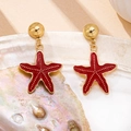 Detailed close-up of golden starfish earrings with red enamel on a decorative shell - Red