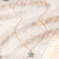 Vue détaillée du pendentif étoile de mer avec émail bleu et chaîne dorée sur sable blanc - Bleu sarcelle