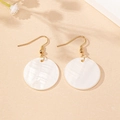 Detail of shell disk drop earrings with gold hooks on a minimalist textured surface - White