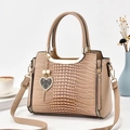 Bolso beige con grabado de cocodrilo, asa metálica y colgante de corazón en mesa blanca - Caqui