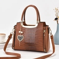 Detalle de bolso marrón con diseño de cocodrilo, asa metálica y colgante de corazón - Amarillo Tierra