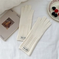 Close-up of cream knitted gloves with rib-knit cuffs and fabric label on a white bed sheet - Cream