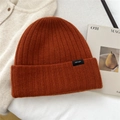 Macro detail of burnt orange 100% wool knitted beanie featuring a black square brand label - Orange