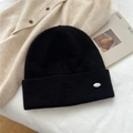 Close-up of black knitted beanie texture with silver oval metal badge on folded brim - One Size / Black