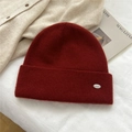 Detail of soft dark red knit beanie with silver metal emblem on white fabric background - One Size / Red With Oval Metal Tag