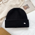 Detalle del gorro de punto acanalado negro con placa metálica sobre cama acogedora - Talla única / Negro