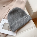Detalle del gorro de punto acanalado gris con etiqueta metálica sobre fondo beige - Talla única / Gris claro