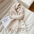 Detail view of a creamy white knitted scarf with soft tassels and metallic logo detail - Cream